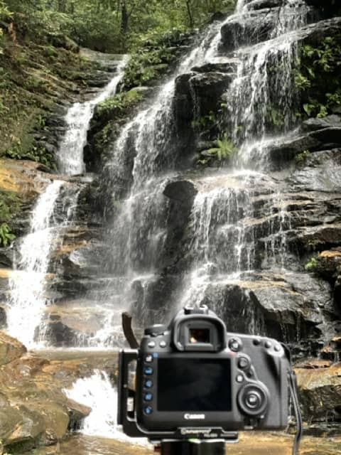 A person holding a camera in front of a waterfall in the Blue Mountains.
