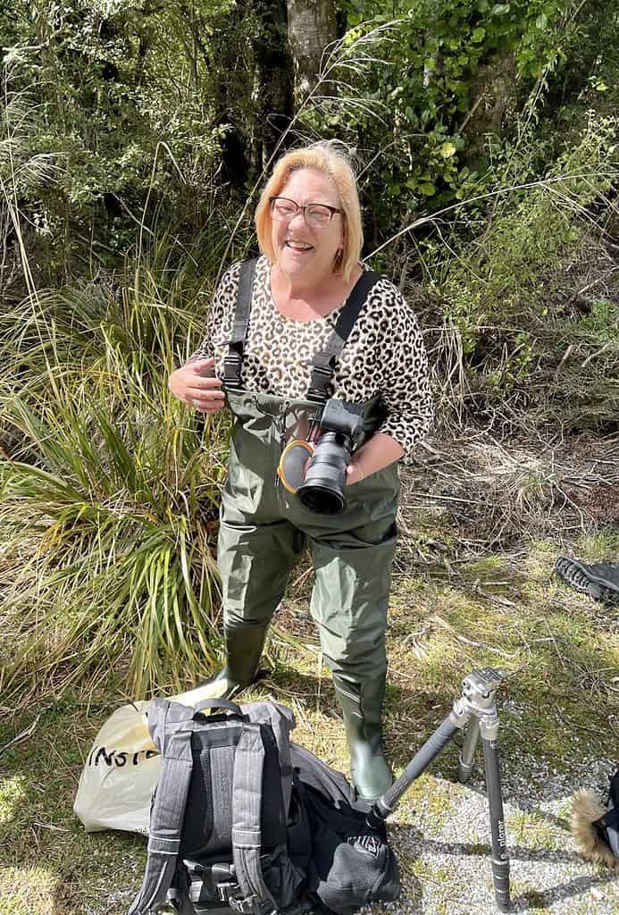 woman wearing fishing waders holding a camera and laughing - solo travel fun