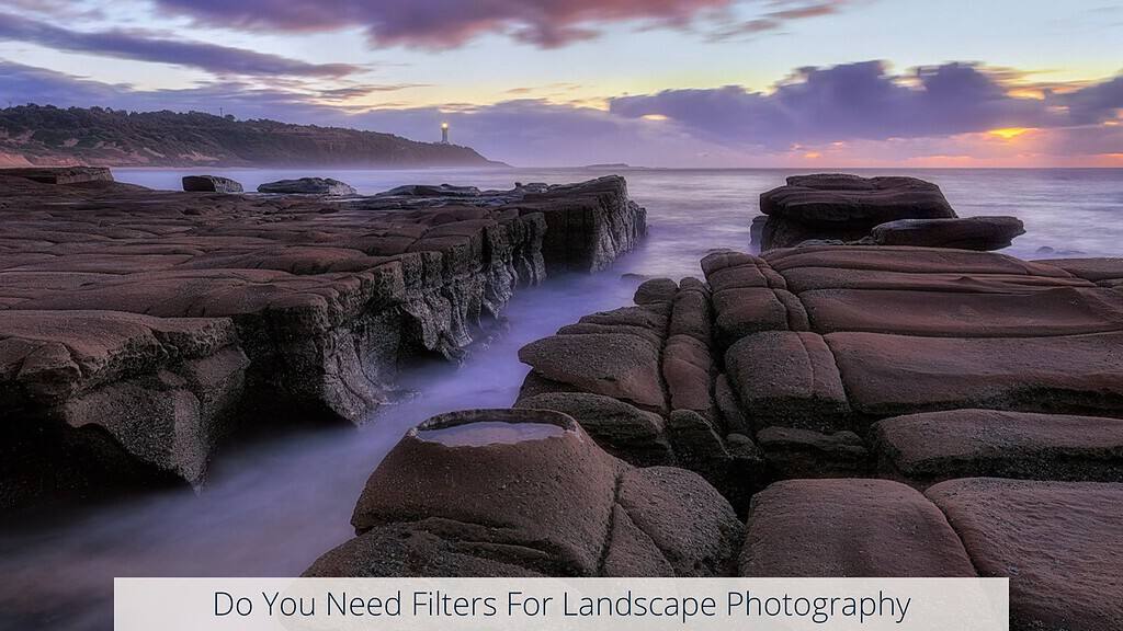 Dramatic coastal landscape at dusk with rugged, layered rock formations leading to the sea and a lighthouse visible in the distance. Text overlay on the image asks 'Do You Need Filters For Landscape Photography,' hinting at the techniques possibly used to enhance the vibrant colors and smooth water effect in this scenic photograph