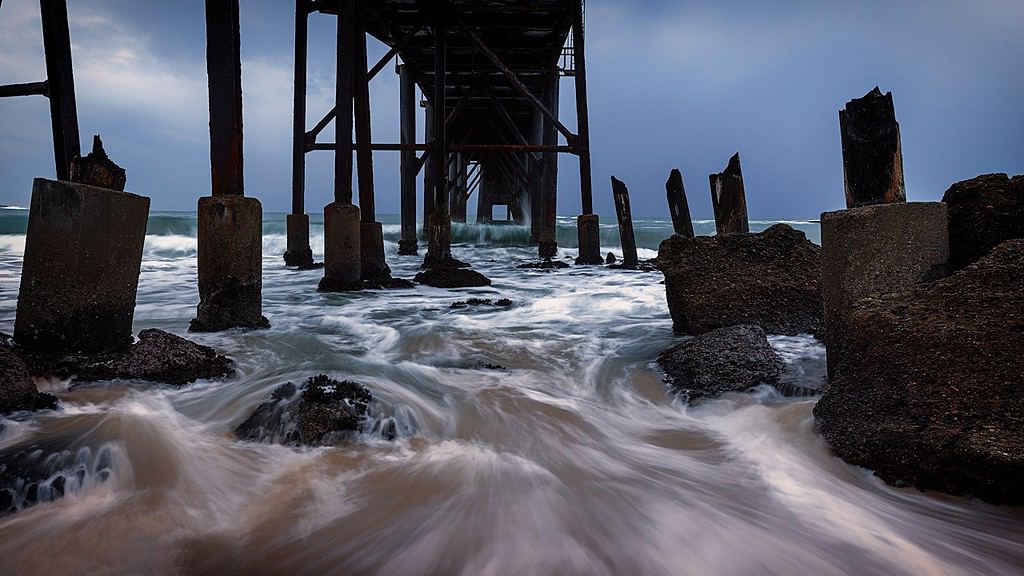Dynamic seascape depicting the powerful ocean waves flowing beneath a weathered pier, illustrating the concept of 'frame within a frame' in landscape photography through the decaying structural elements