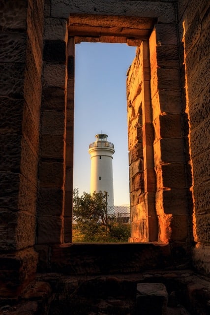 A lighthouse seen through an old stone window, with the warm glow of sunset creating a 'frame within a frame' effect in this serene landscape photo.