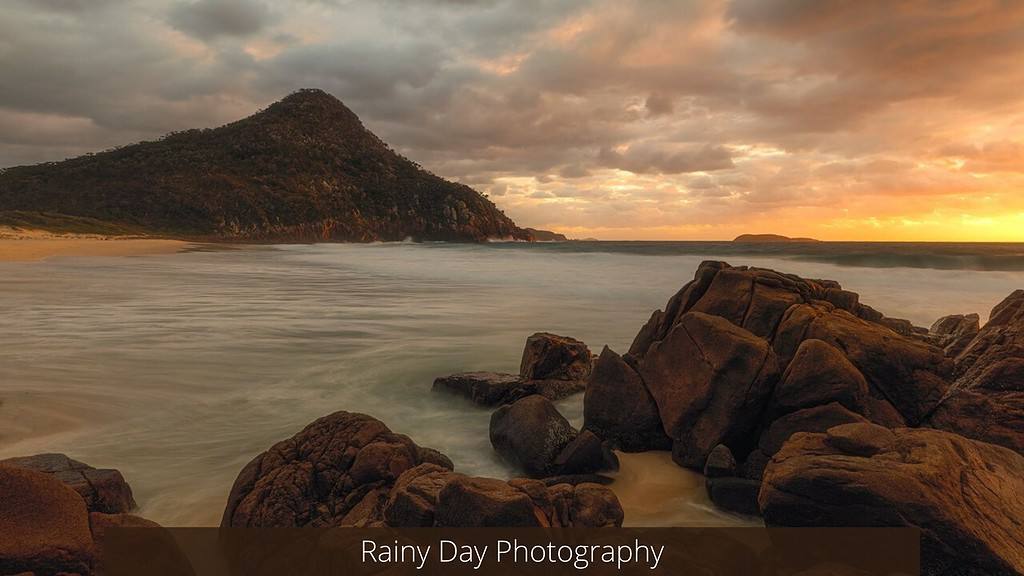 Dramatic seaside landscape captured on a cloudy day, featuring smooth, long-exposure waves caressing a rugged coastline with large brown rocks in the foreground and a dark, forested mountain in the background, under a moody, golden-lit sky with the title "Rainy Day Photography" overlaid at the bottom.
