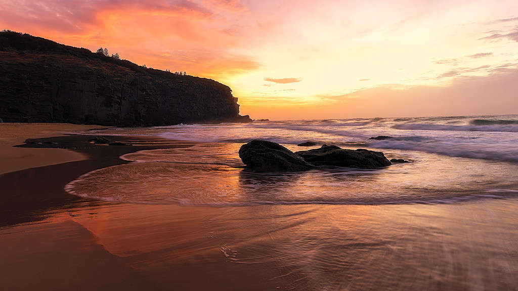 A breathtaking sunrise over a serene beach, with the sky painted in shades of pink and orange, reflecting on the wet sand and gentle waves, bordered by a rugged cliff. Location - Redhead Beach, NSW, Australia
