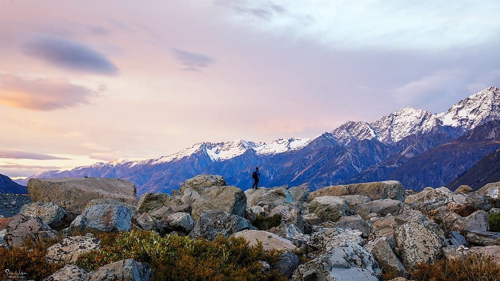 Person in blue standing atop rocky foreground overlooking a majestic mountain range during sunset, with hues of purple and orange in the sky