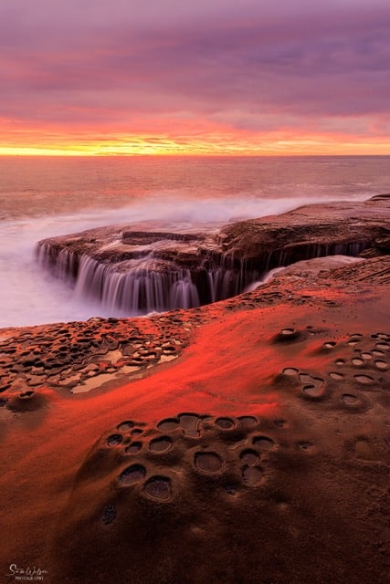 Ocean waves flowing into a natural rock formation creating a waterfall effect, against a backdrop of a sunrise sky