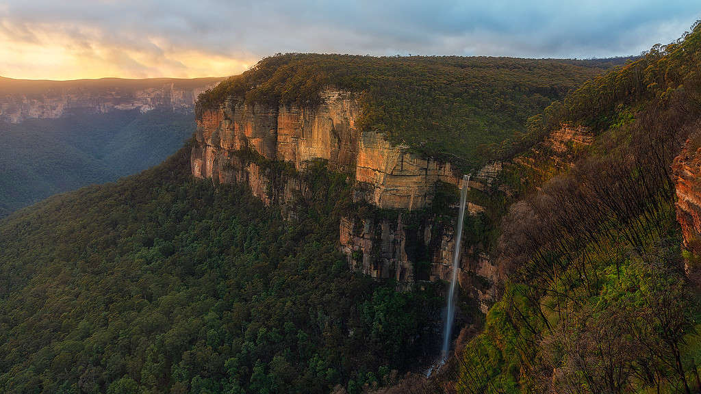 Vast cliffside with a tall waterfall cascading down into lush greenery, as the sun sets casting a warm glow over the scene. Location is the Blue Mountains in Australia