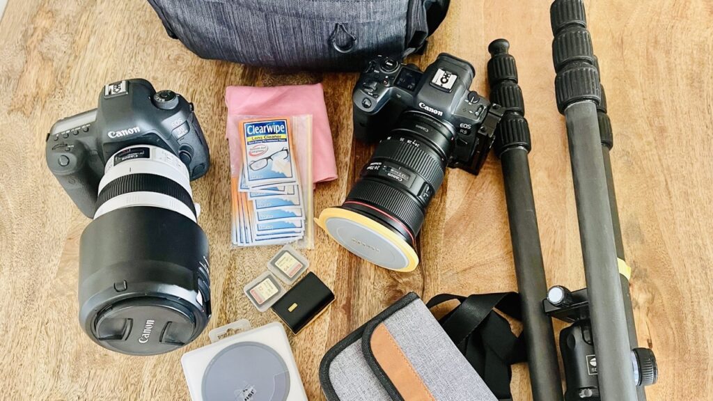 A collection of photography equipment laid out on a wooden floor, including cameras, lenses, memory cards, and a tripod, essential gear for a photographer.