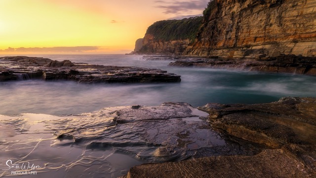 Landscape photography of a beach at sunrise with smooth waters washing over rocks and a vibrant sky, demonstrating the importance of planning landscape photography to capture such tranquility