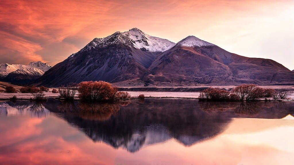 A vibrant landscape reflection photo featuring snow-capped mountains at twilight with the sky's pink and purple hues mirrored in the still water, exemplifying the dramatic effect of clouds and lighting in landscape photography