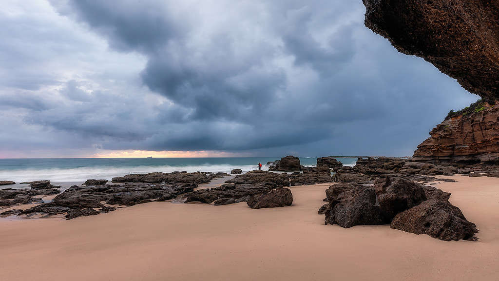 A dramatic coastal landscape under a brooding, textured sky with a person in red adding a scale and human element, illustrating the impact of cloud formations on the mood of landscape photography